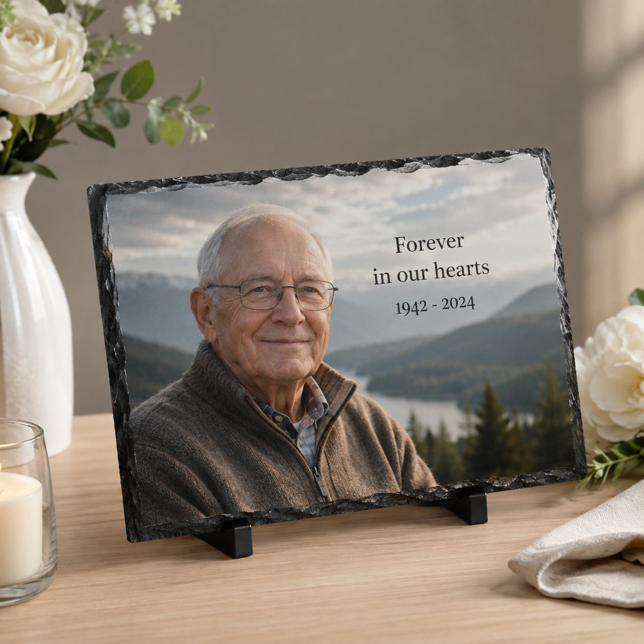 Memorial slate of a smiling man reading “Forever in our hearts 1942–2024”, set among white roses and a candle